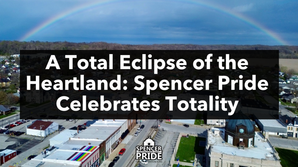 A rainbow stretches across a view of downtown Spencer, Indiana. In the foreground, the Spencer Pride commUnity center appears with a large rooftop rainbow flag.